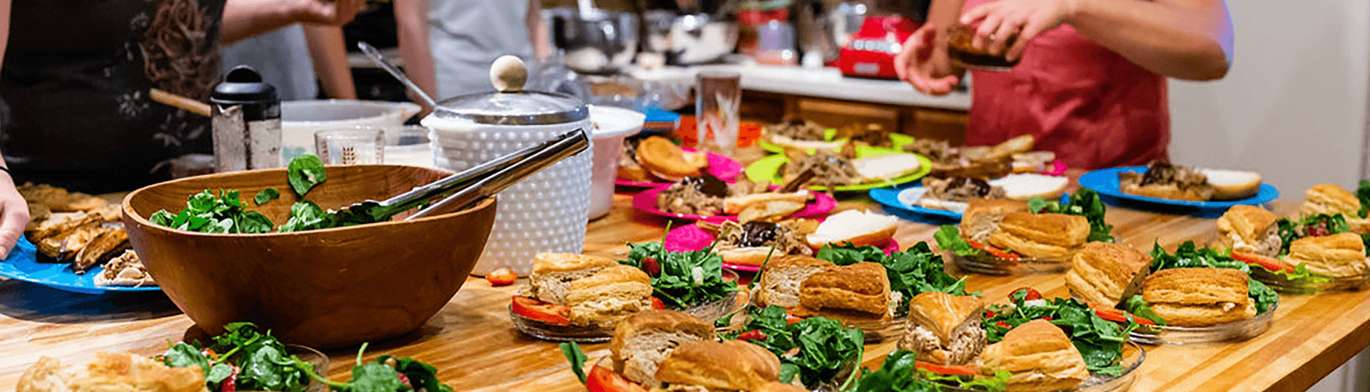 A wooden table is filled with plates of sandwiches and fresh salad, surrounded by people preparing food.