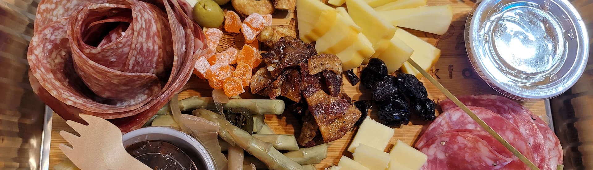 A wooden platter featuring an assortment of cured meats, cheeses, olives, nuts, dried fruits, and pickled vegetables.
