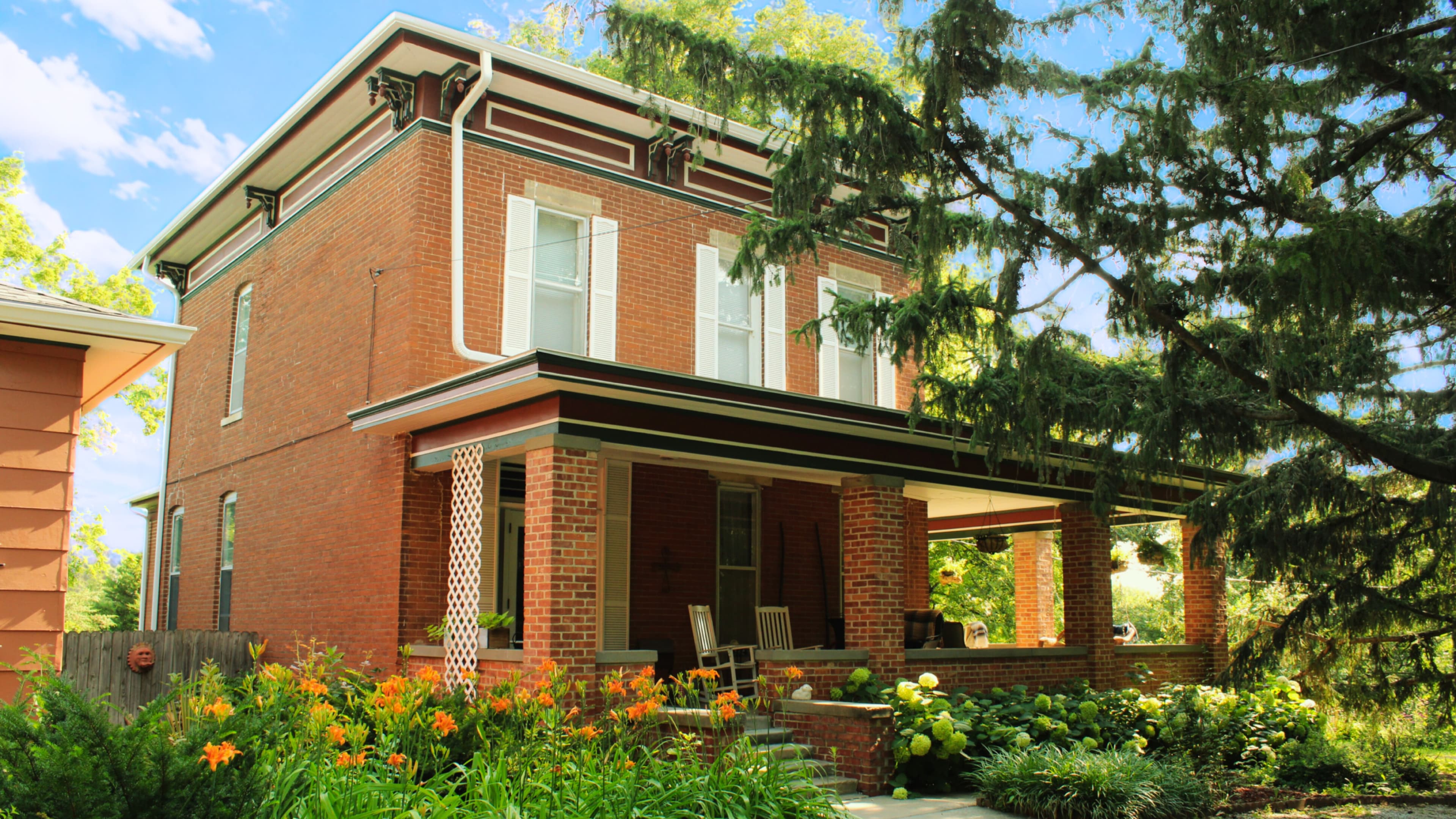 A red brick house with a porch surrounded by greenery and colorful flowers.