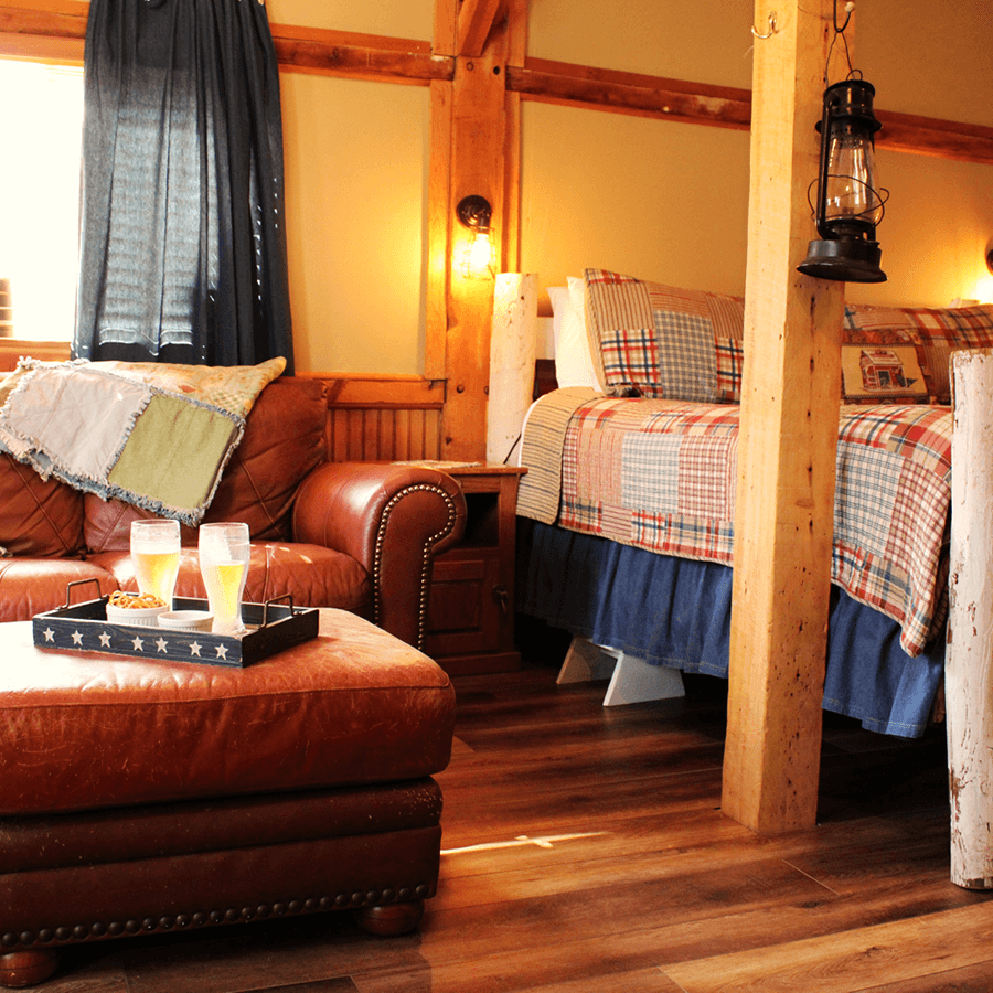 Bedroom with wood floors, couch, padded foot stool with tray and 2 glasses of bubbly, and american quilt on bed