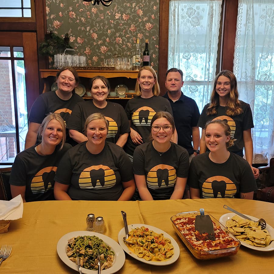 A group of nine people in matching t-shirts poses together at a dining table filled with food.